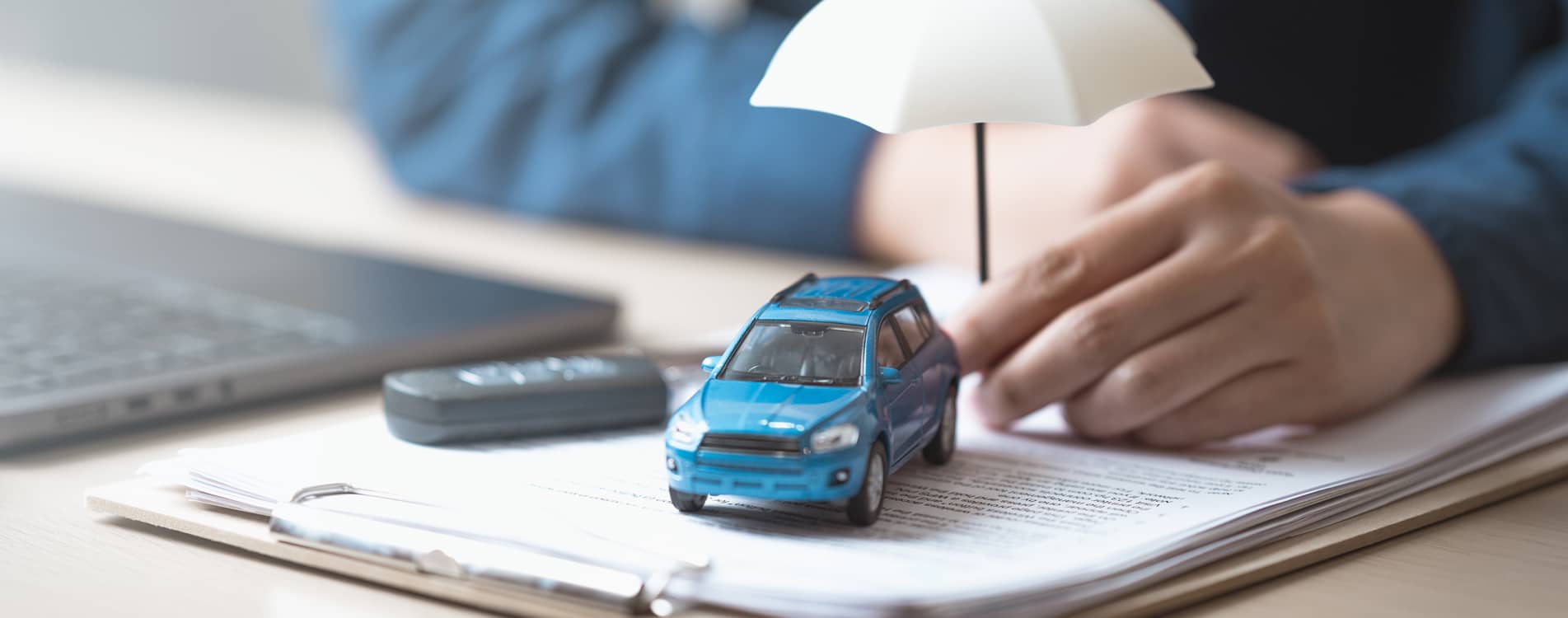 Person holding a small umbrella next to a miniature car, paperwork and car keys.
